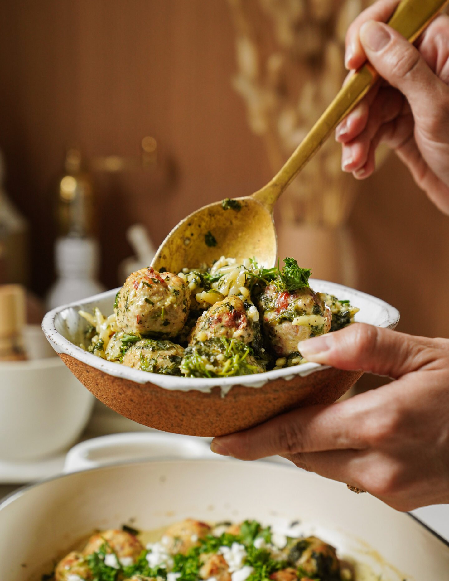 A person serves a bowl of orzo pasta with meatballs and greens using a large spoon, held above a white pot with similar food. The dish looks fresh and garnished with herbs.