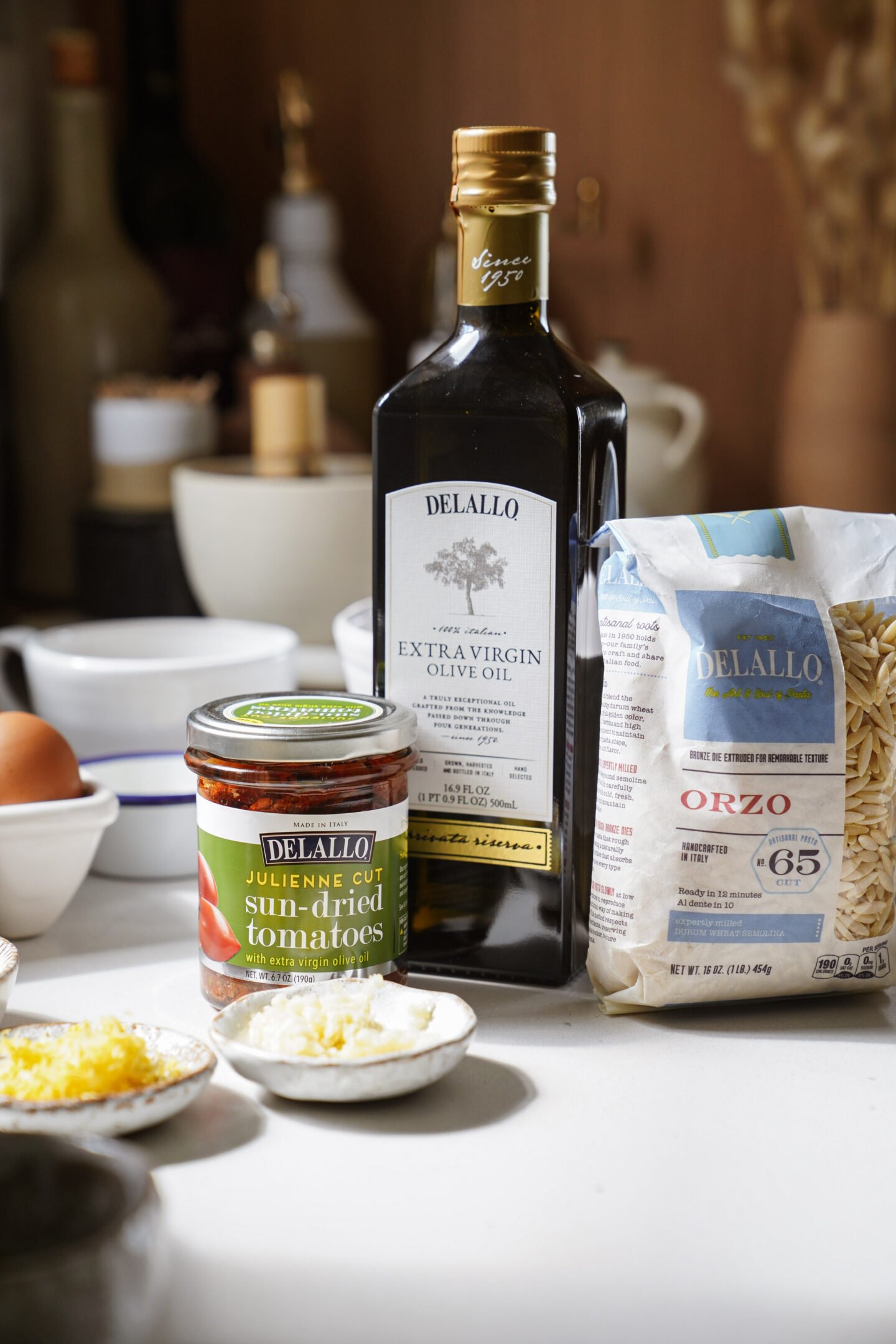 A bottle of DeLallo extra virgin olive oil, a package of DeLallo orzo pasta, and a jar of DeLallo sun-dried tomatoes are arranged on a kitchen counter with bowls and ingredients nearby.