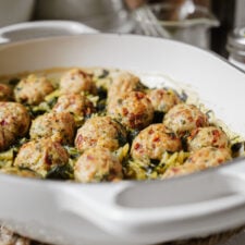 Close-up of a white baking dish filled with golden-brown meatballs on a bed of greens and orzo pasta, freshly baked and ready to serve.