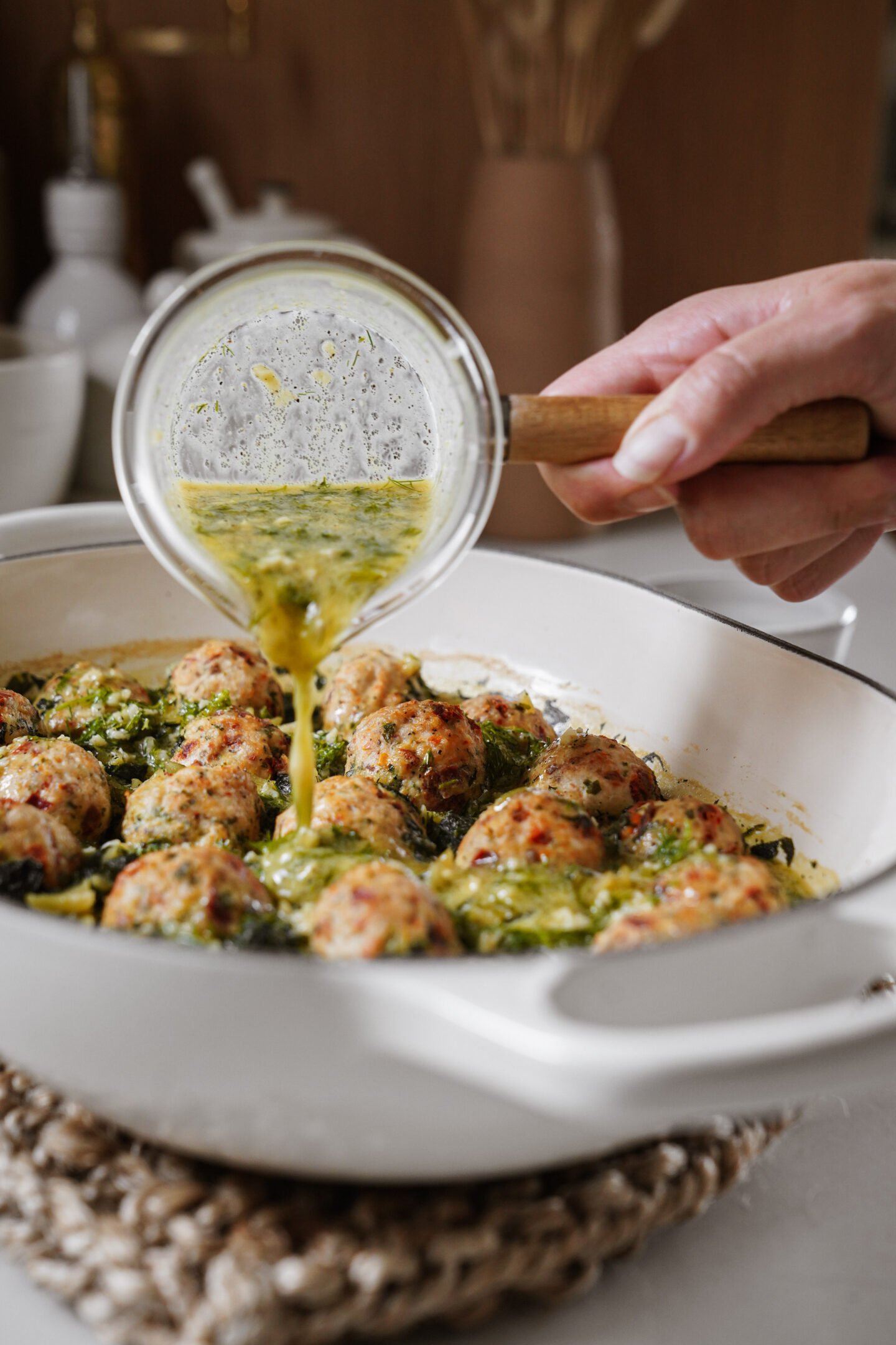 A hand pours a herbed sauce from a small glass bowl over baked meatballs in a white casserole dish, sitting on a woven trivet on a kitchen counter.