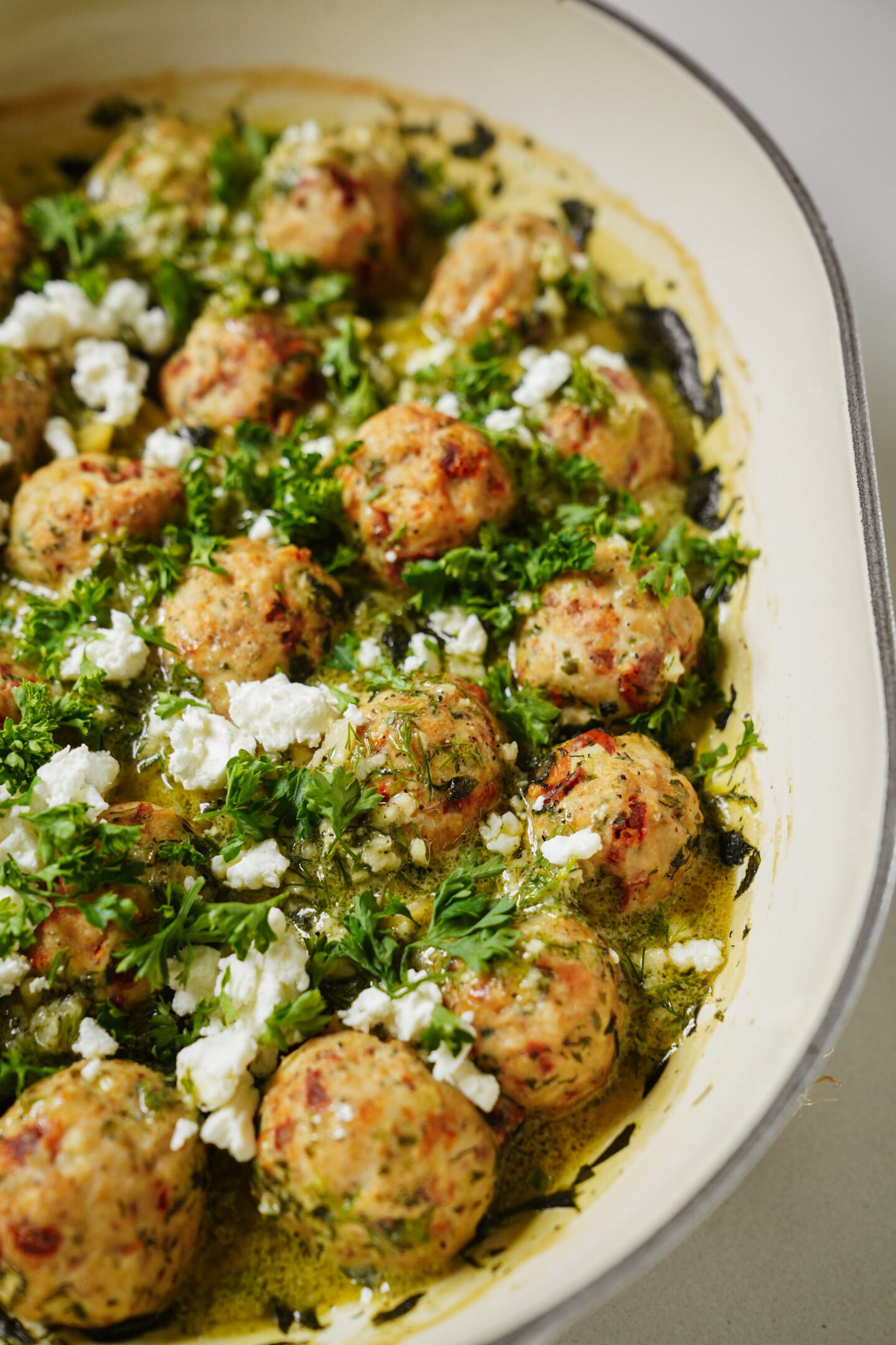 A close-up of a baking dish filled with chicken meatballs in a creamy herb sauce, topped with crumbled feta cheese and fresh chopped parsley.