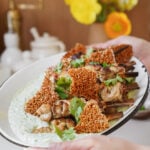A person holds a platter with grilled chicken skewers, garnished with cilantro, placed over a creamy green sauce and topped with crunchy sesame crisps. Bright yellow flowers and kitchen items are blurred in the background.