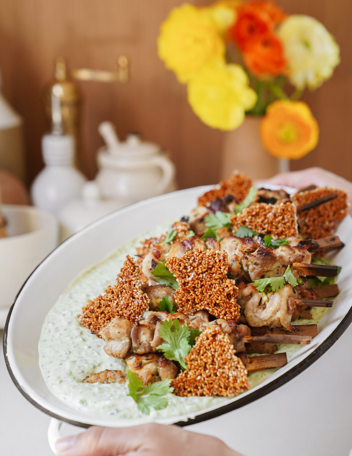 A platter of grilled chicken skewers with crunchy sesame crisps, garnished with fresh cilantro, served over a creamy green sauce. A bright bouquet of orange and yellow flowers is blurred in the background.