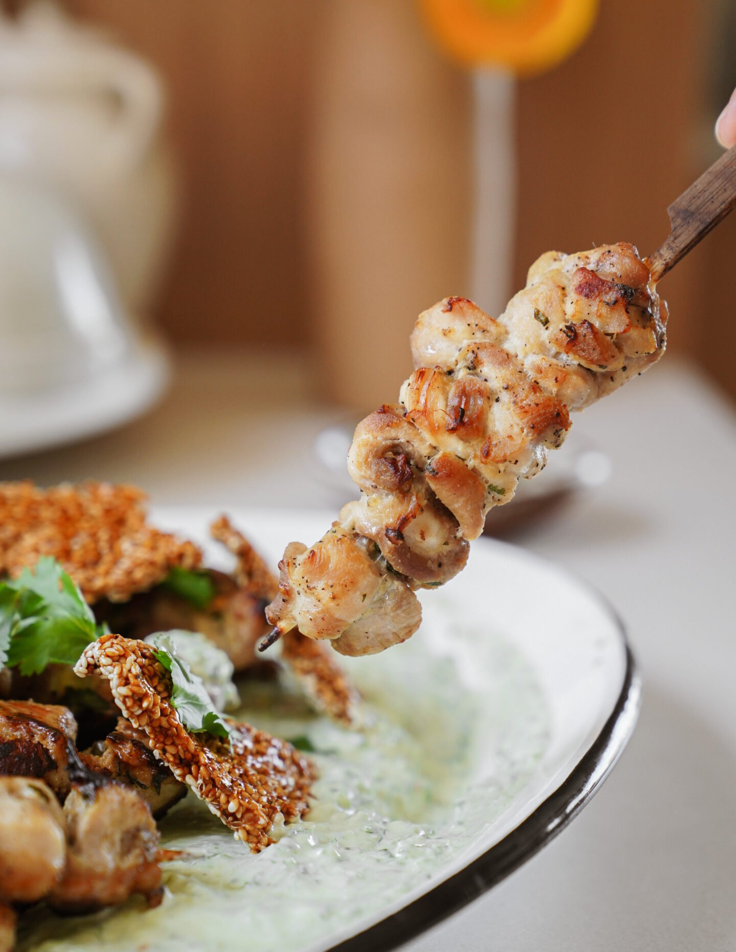 A close-up of a grilled chicken skewer being held above a plate with a green sauce and sesame crisps, garnished with fresh herbs.