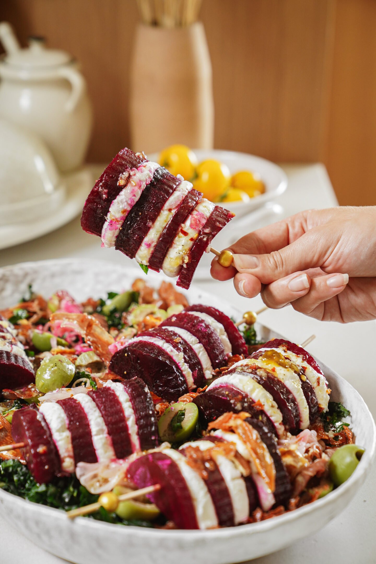 A hand holds a skewer of sliced beets layered with creamy cheese above a platter of salad featuring greens, olives, and more beet and cheese skewers.