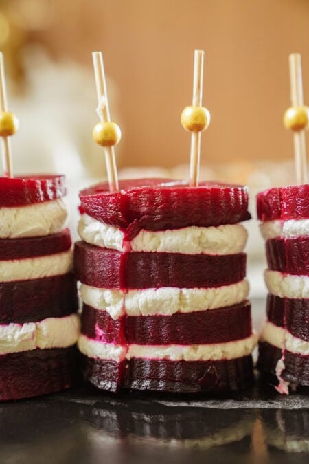 Stacked slices of beetroot and goat cheese alternately layered, held together with decorative skewers topped with gold beads, arranged on a dark surface.