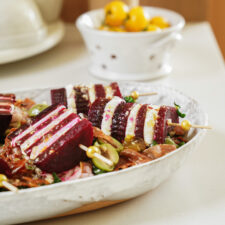 A close-up of skewers with alternating layers of beetroot and cheese, arranged on a white platter with olives and greens. In the background, there is a bowl of yellow tomatoes.