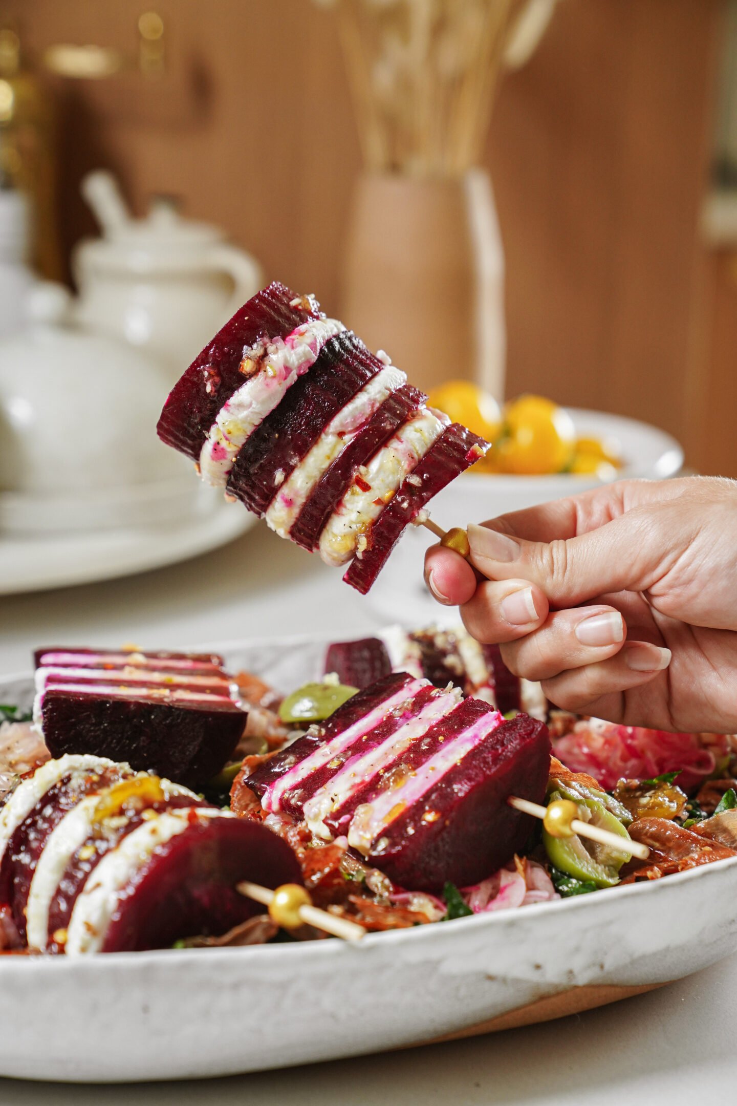 A hand holds a skewer with stacked layers of beetroot, cream cheese, and herbs. More skewers with the same layered beetroot and cheese are arranged on a white serving dish in the background.