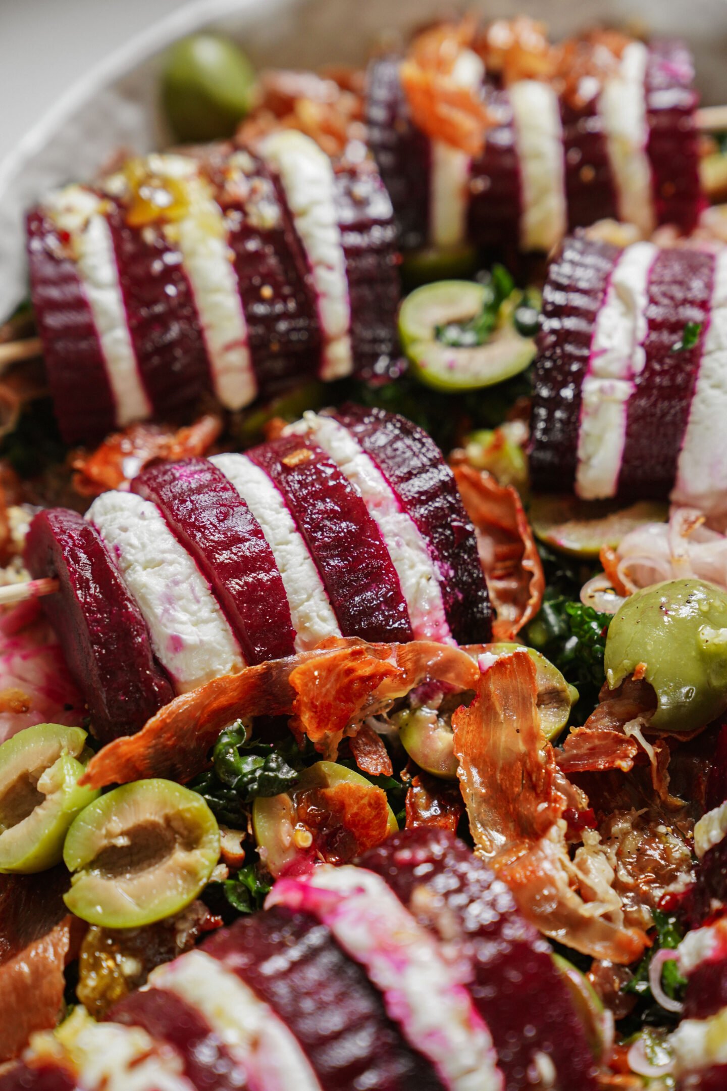 Sliced beetroot and goat cheese stacks arranged on a salad with green olives, crispy prosciutto, and greens, drizzled with dressing.