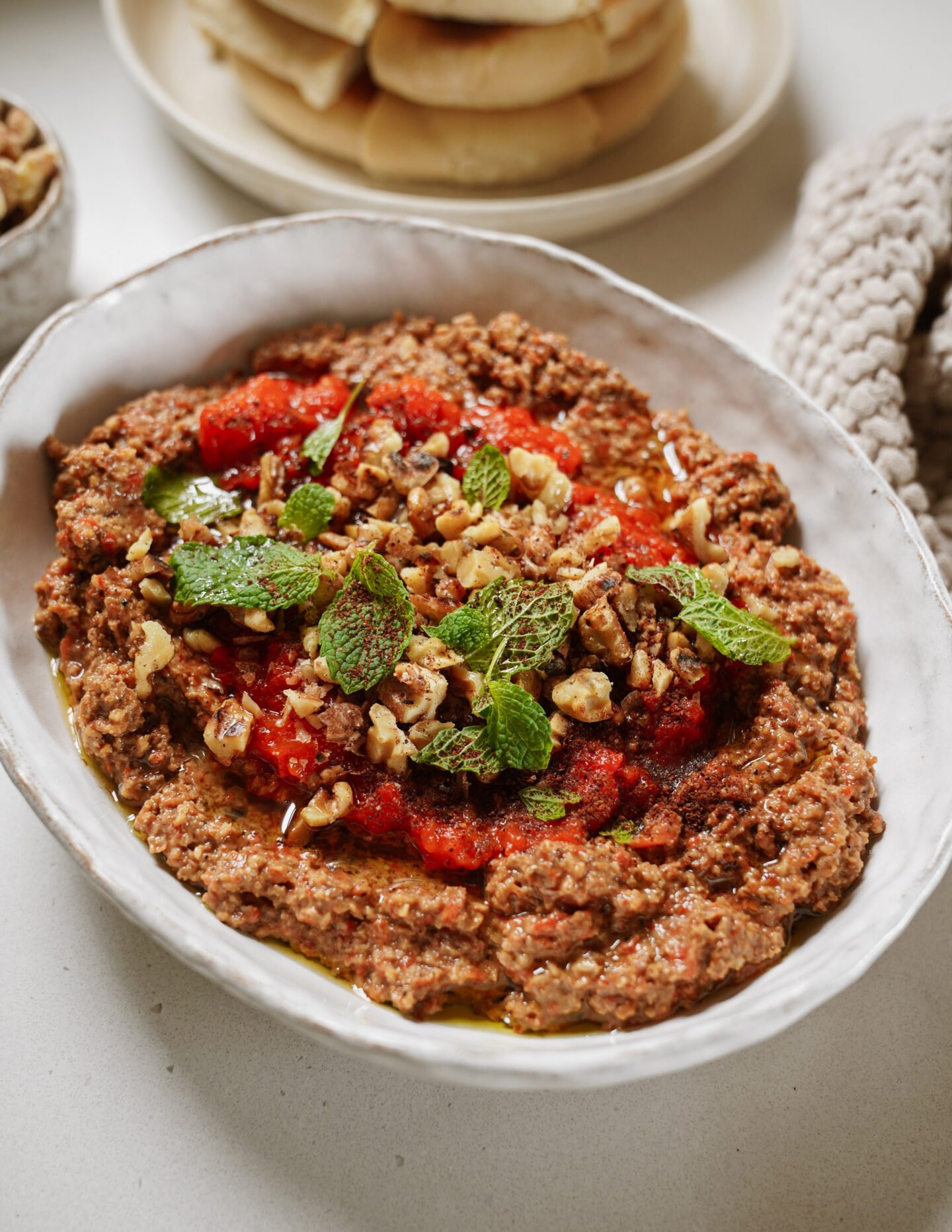 A white oval bowl filled with a chunky dip topped with chopped walnuts, fresh mint leaves, and a drizzle of oil, with pita bread and a textured cloth in the background.