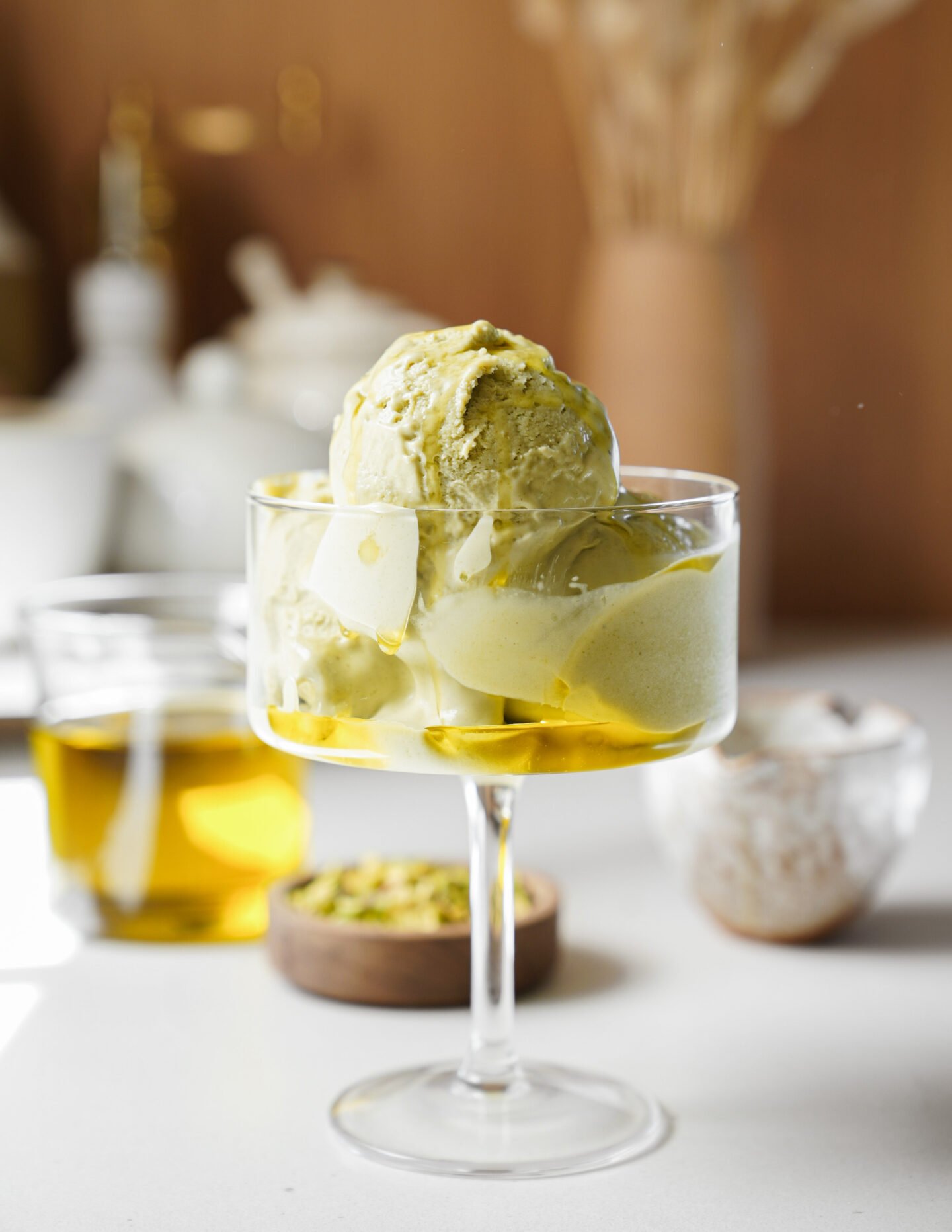 A glass dish filled with green pistachio ice cream drizzled with olive oil, set on a white surface with a small glass of olive oil, a bowl of pistachios, and blurred background objects.