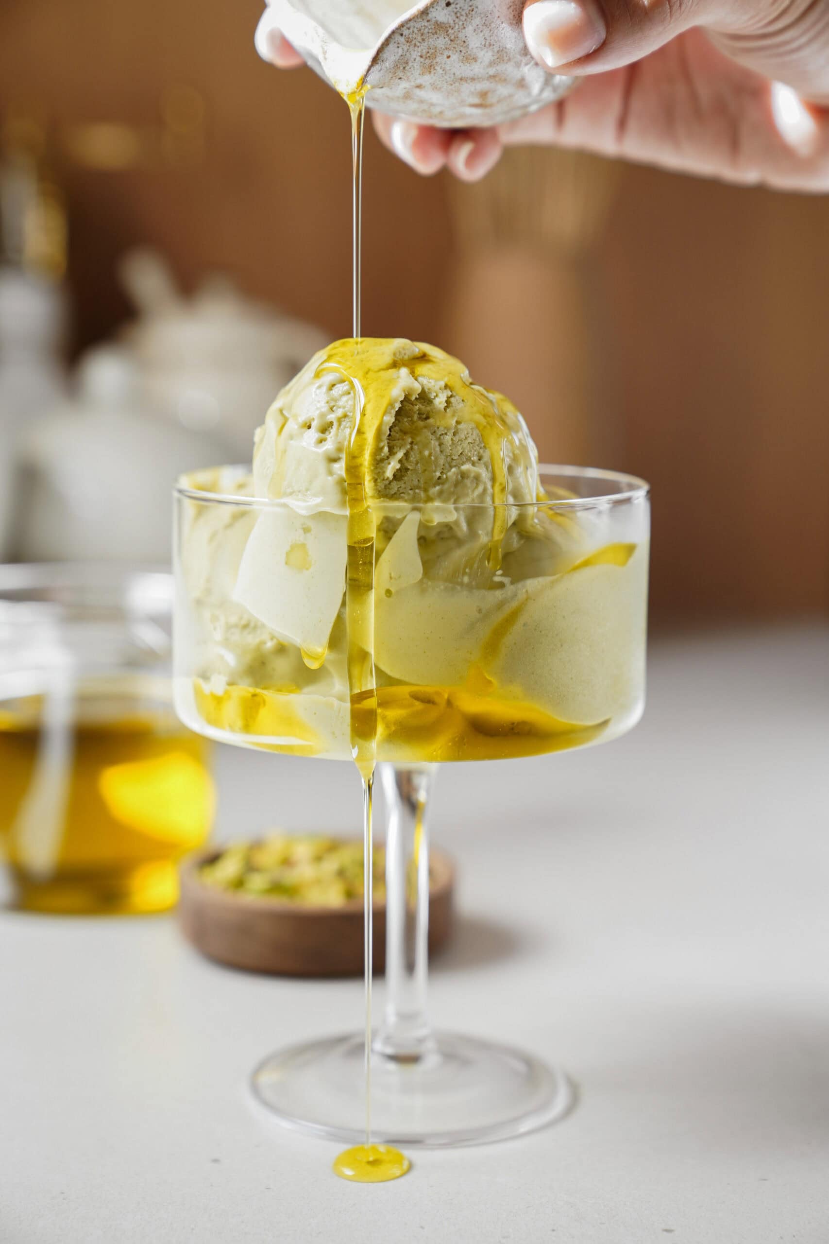 A hand pours golden olive oil over two scoops of pistachio ice cream served in a glass dish, with a jar of olive oil and a small bowl of pistachios in the blurred background.