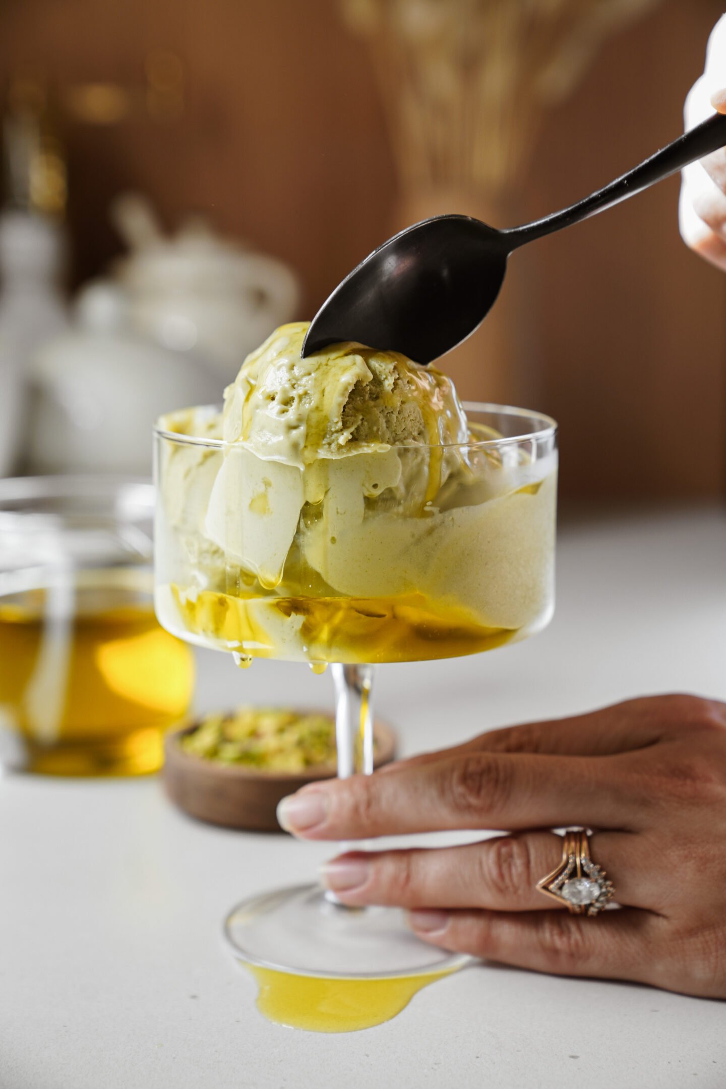 A hand holds a glass dish with a scoop of pistachio ice cream as honey is drizzled on top with a spoon. A jar of honey and a bowl of pistachios are blurred in the background.