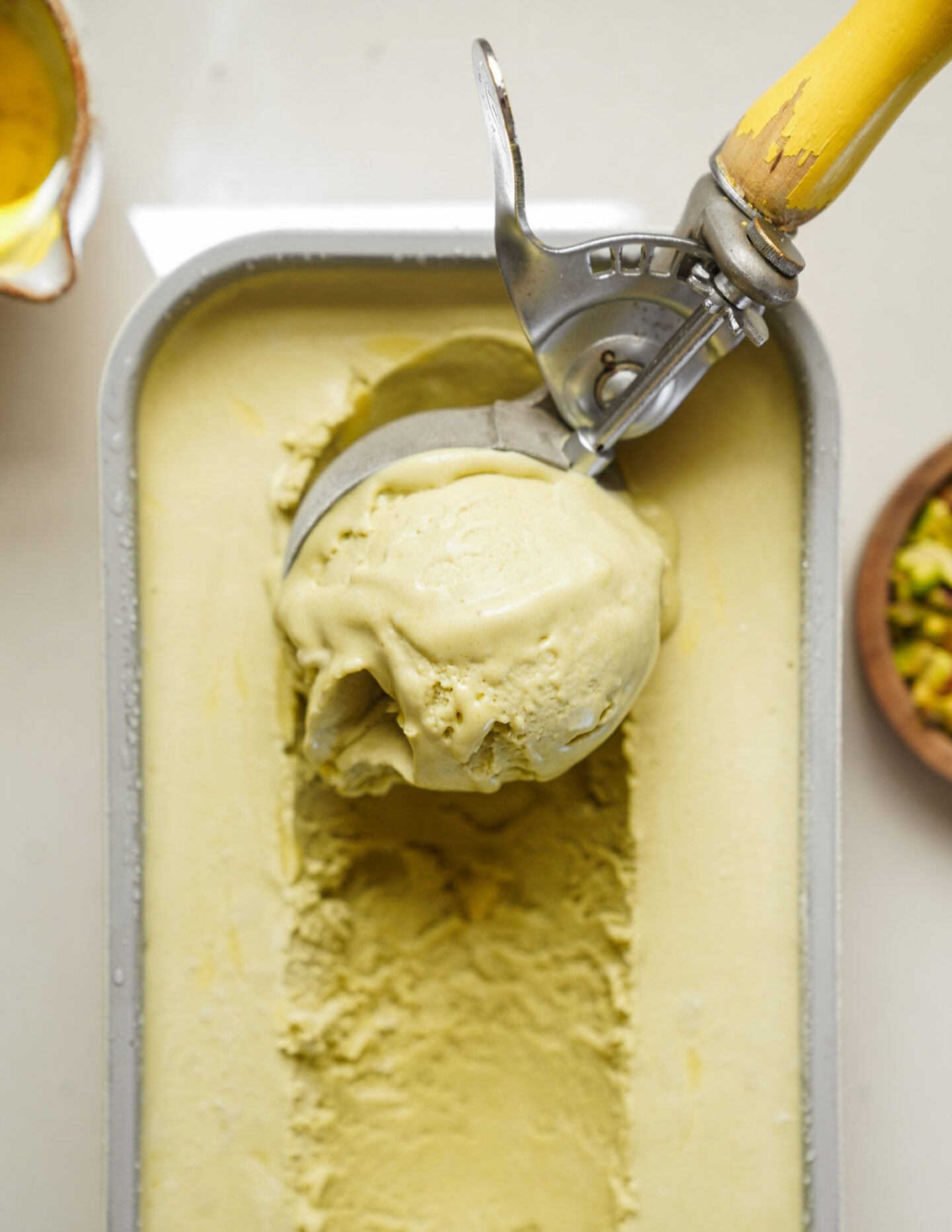 A metal ice cream scoop with a yellow handle is lifting a scoop of pale green pistachio ice cream from a rectangular metal container.
