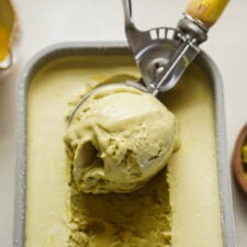 A close-up of a metal ice cream scoop lifting a large serving of pistachio ice cream from a rectangular container. The ice cream is pale green and creamy in texture.