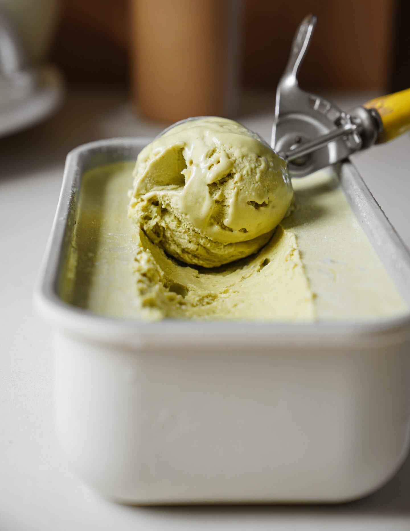 A close-up of a metal ice cream container filled with pale green ice cream, with a scoop being lifted out using a metal ice cream scoop with a yellow handle.