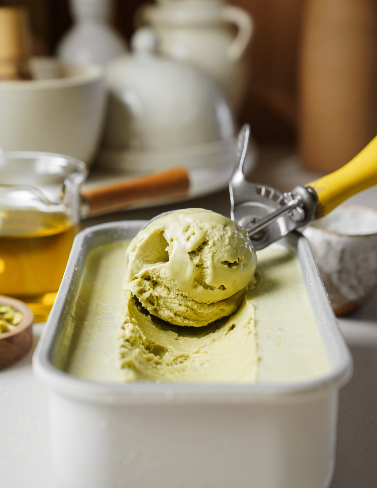 A metal container of green pistachio ice cream with a yellow-handled scooper holding a single scoop. Blurred kitchen items and ingredients are visible in the background.