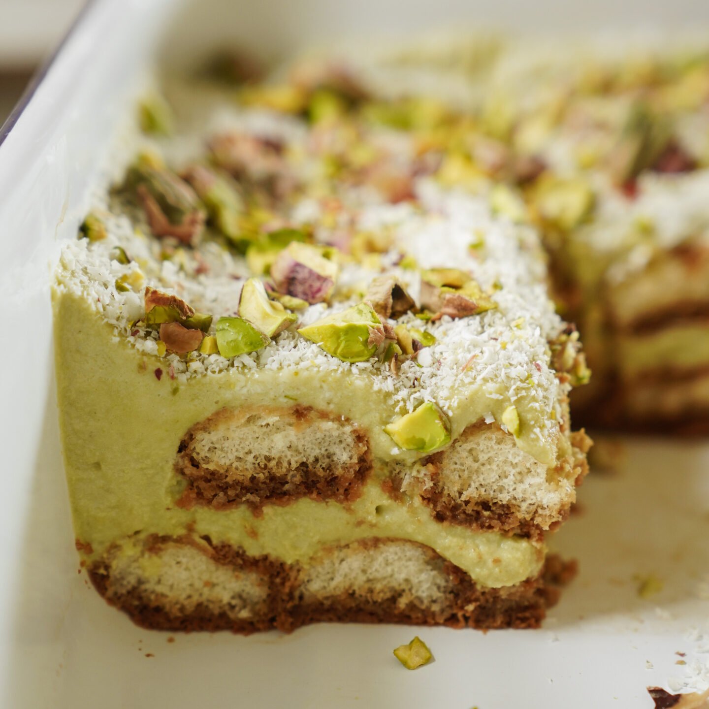 A close-up of a sliced pistachio tiramisu in a white dish, showing layers of soaked ladyfingers, green pistachio cream, and chopped pistachios sprinkled on top.