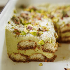 A close-up of a sliced pistachio tiramisu in a white dish, showing layers of soaked ladyfingers, green pistachio cream, and chopped pistachios sprinkled on top.
