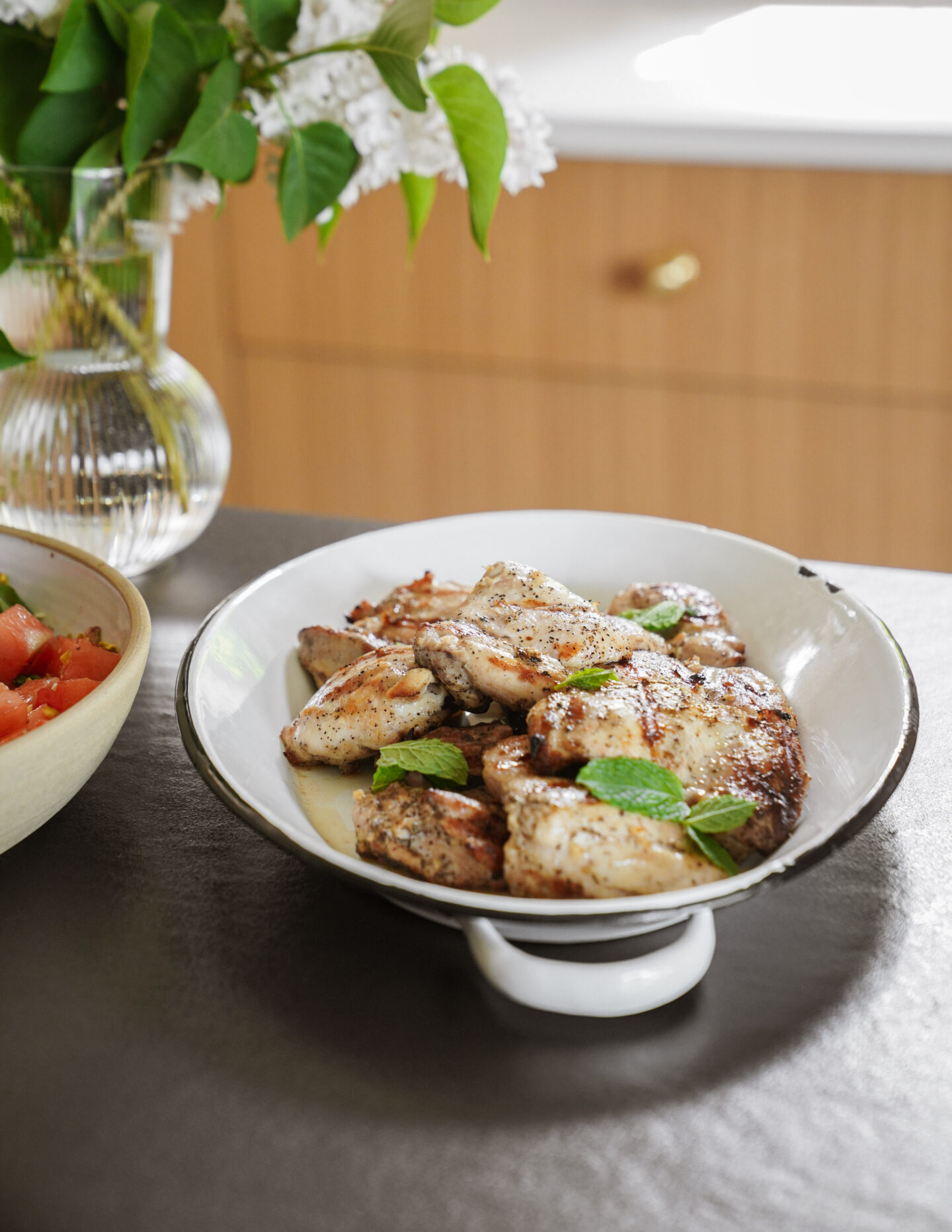 A white dish filled with grilled chicken pieces garnished with fresh mint leaves, sitting on a dark countertop. A bowl of salad and a vase of white flowers are visible in the background.