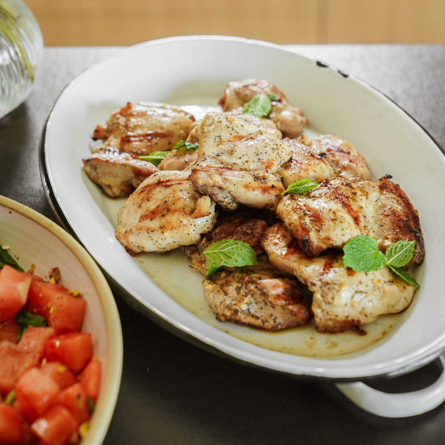 Grilled chicken pieces garnished with fresh mint leaves are served on a white oval platter. A side dish with diced tomatoes and greens is partially visible beside the platter.