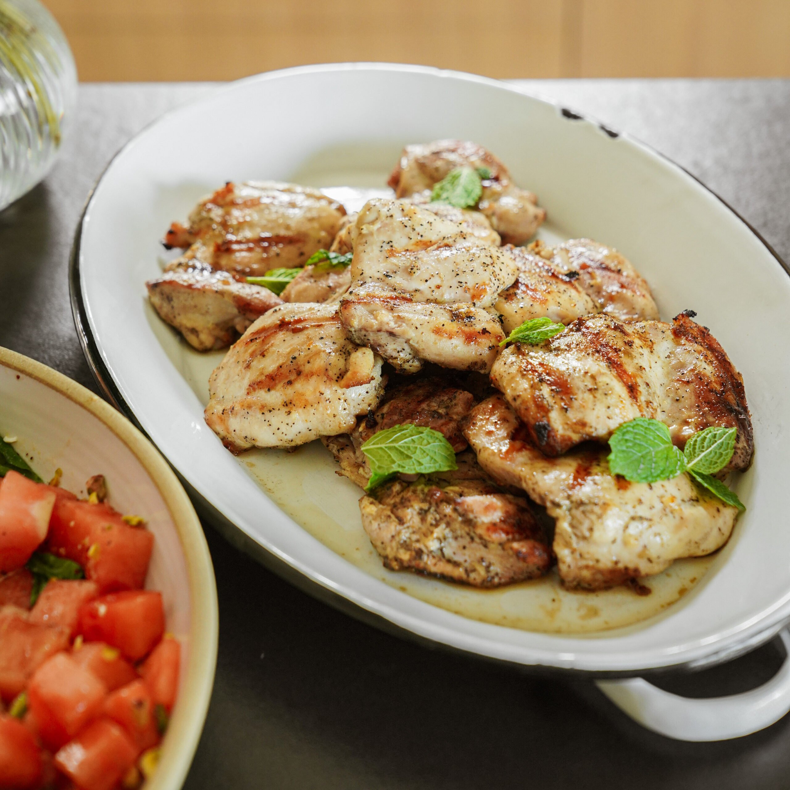 Grilled chicken pieces garnished with fresh mint leaves are served on a white oval platter. A side dish with diced tomatoes and greens is partially visible beside the platter.