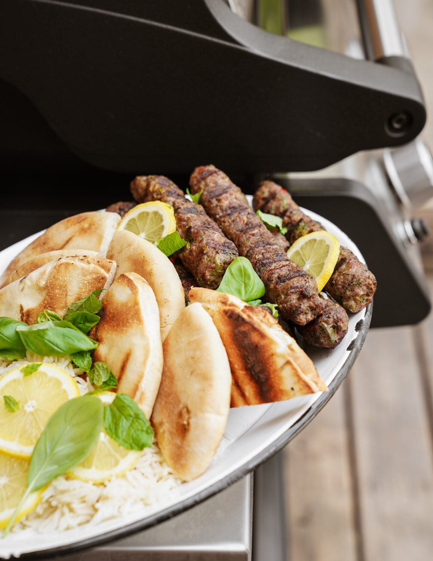 A plate with grilled pita bread, skewered kebabs, white rice, lemon slices, and fresh basil leaves, held near a barbecue grill.