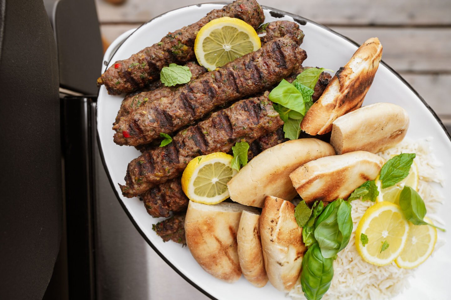 A platter with grilled kebabs, sliced pita bread, rice, fresh basil leaves, and lemon slices, arranged neatly on a white oval plate.