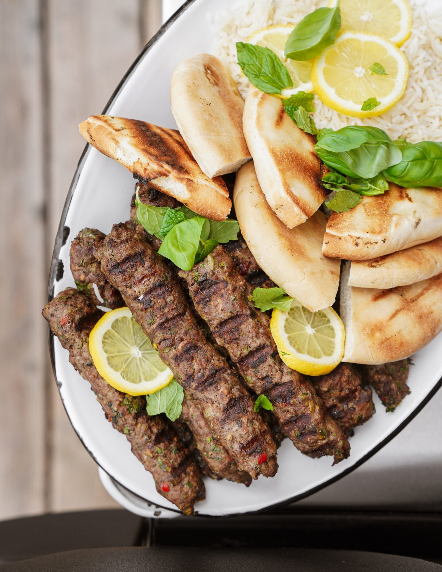Oval plate with grilled kebabs garnished with lemon slices and basil, served with pita bread and white rice topped with lemon and basil, on a wooden surface.