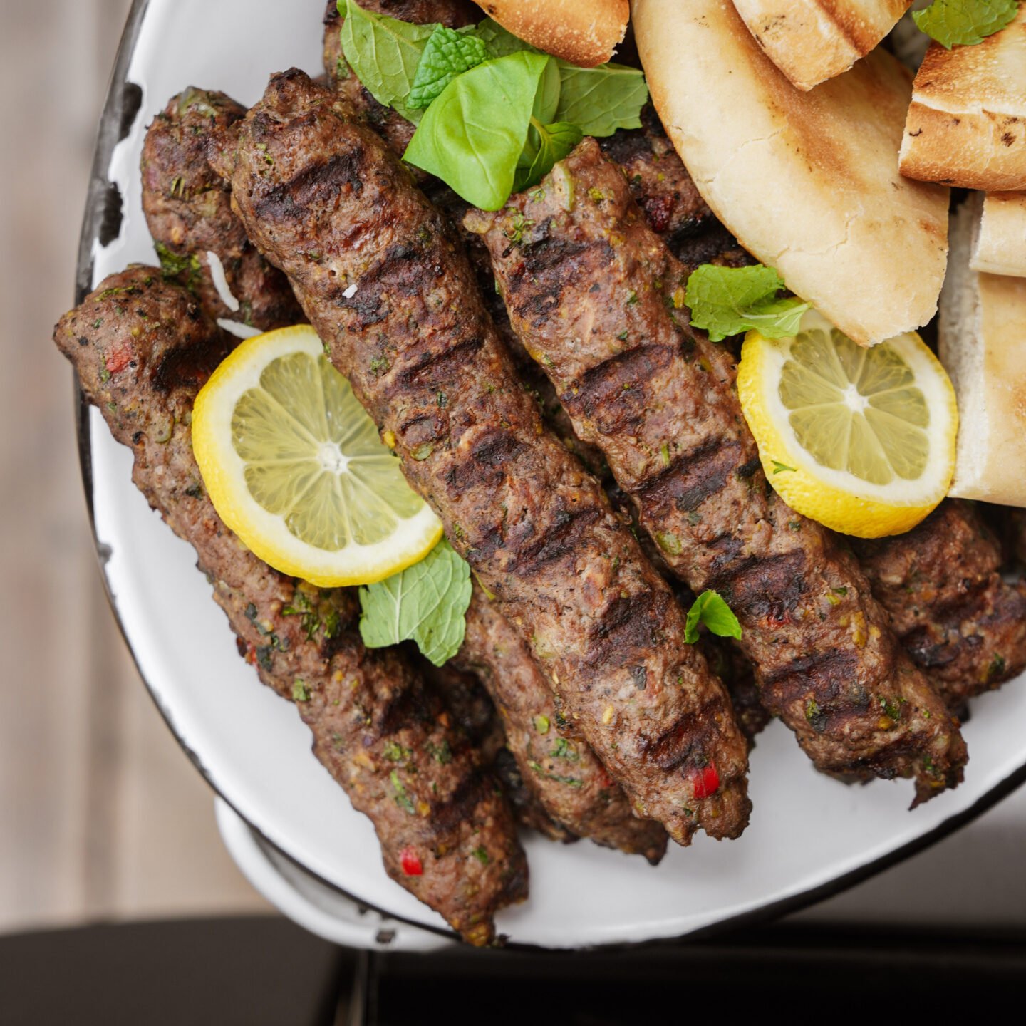 Grilled kebabs garnished with lemon slices and fresh herbs are served on a plate alongside pieces of pita bread.