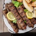 Grilled kebabs garnished with lemon slices and fresh herbs are served on a plate alongside pieces of pita bread.