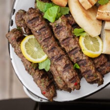 Grilled kebabs garnished with lemon slices and fresh herbs are served on a plate alongside pieces of pita bread.