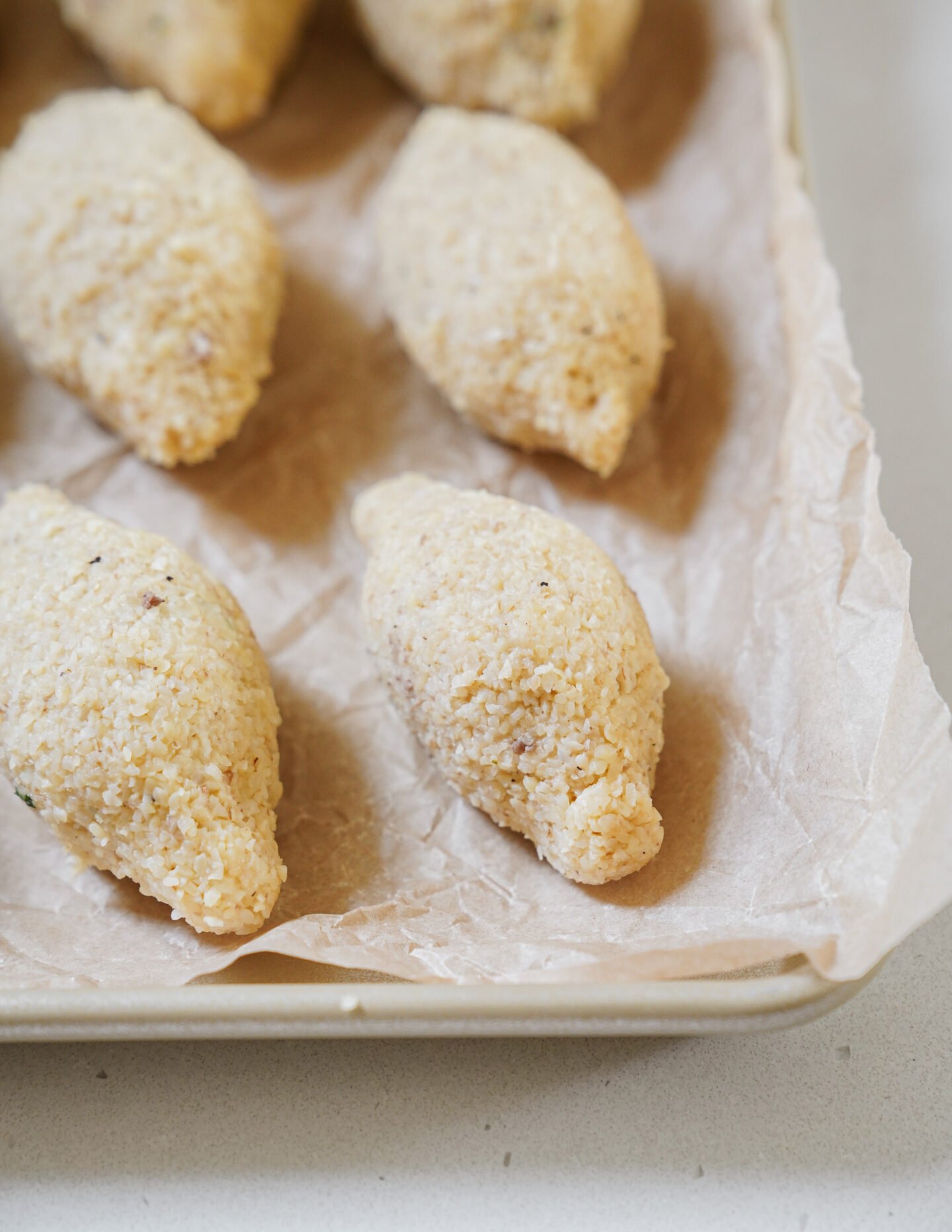 A close-up of several uncooked kibbeh, oval-shaped bulgur wheat and meat dumplings, arranged on a parchment-lined baking tray.
