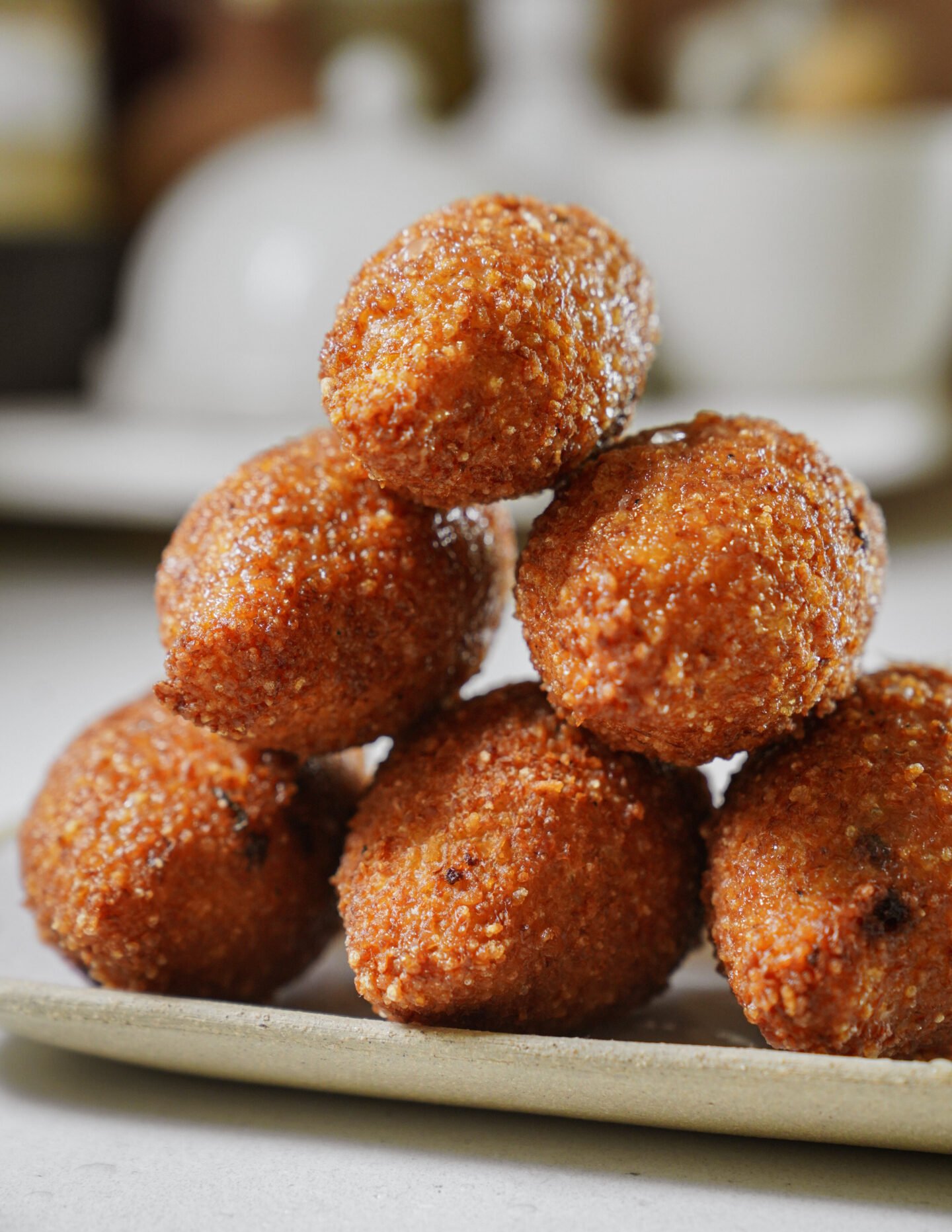 A close-up of six golden-brown, crispy fried balls stacked in a pyramid on a rectangular plate, with a blurred background.