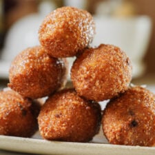 A close-up of six golden-brown, crispy hush puppies stacked in a pyramid on a plate, with a blurred background.