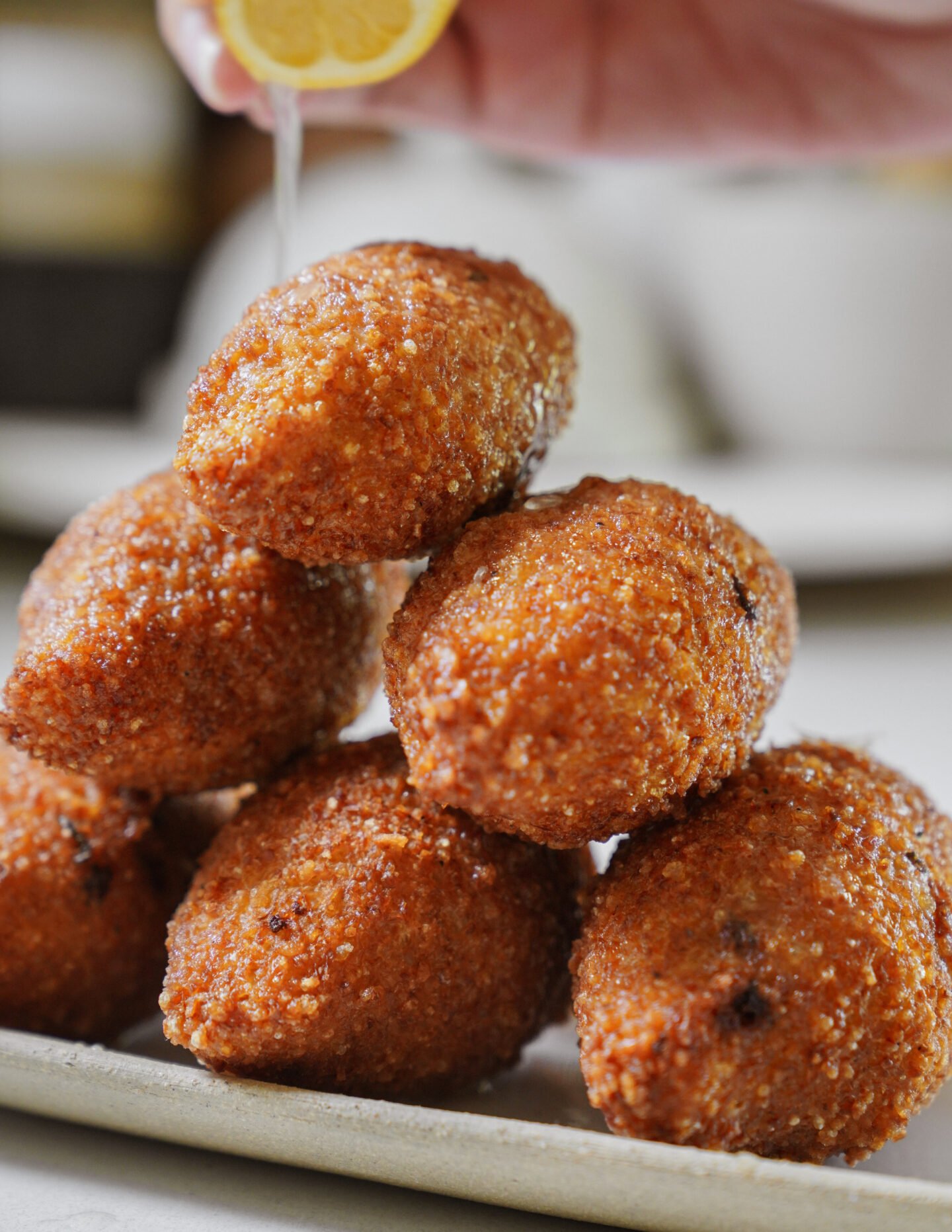 A close-up of a stack of golden, crispy kibbeh on a plate, with a hand squeezing fresh lemon juice over them.