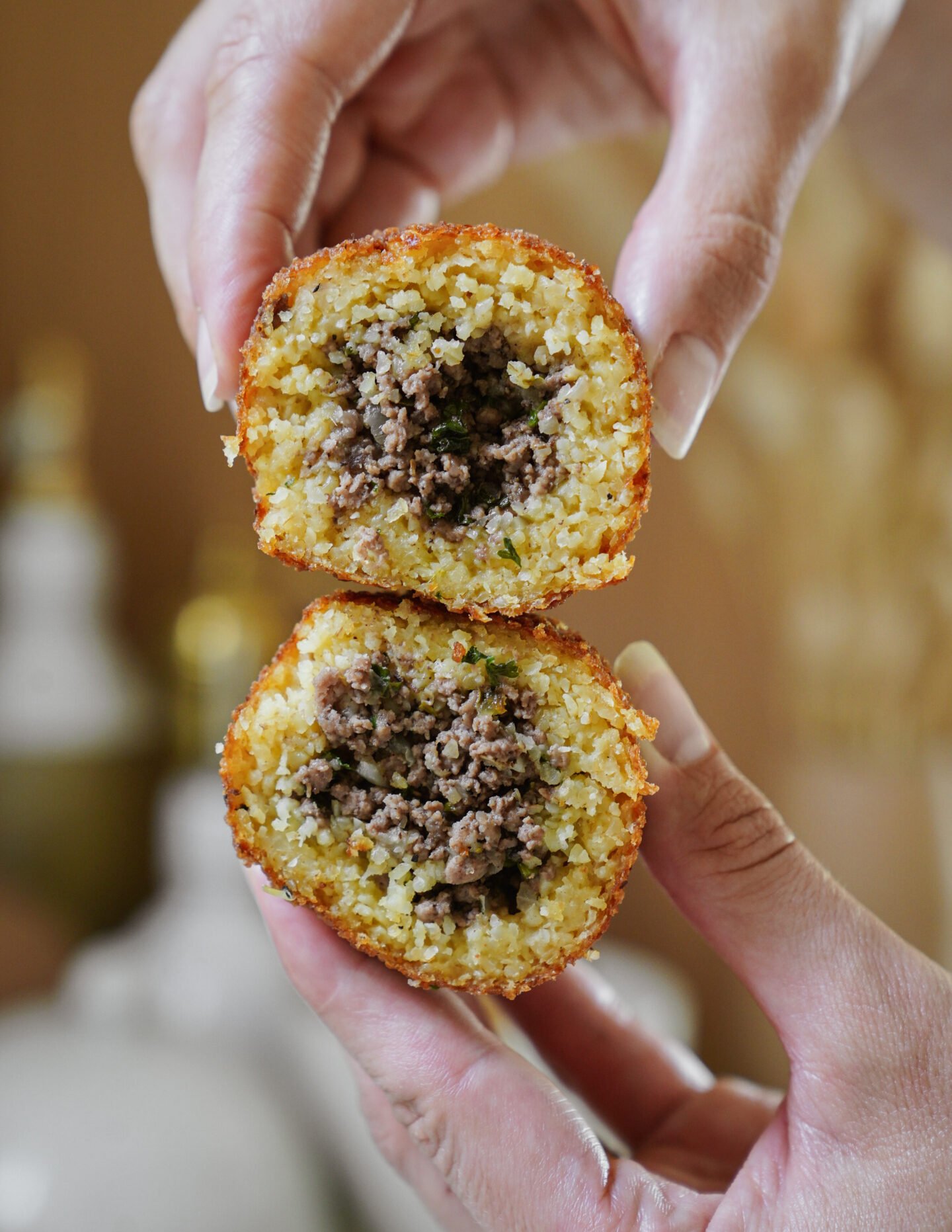 A close-up of two hands holding a golden, crispy ball cut in half, revealing a savory ground meat filling inside. The texture of the outer shell looks crunchy, while the inside appears moist and flavorful.