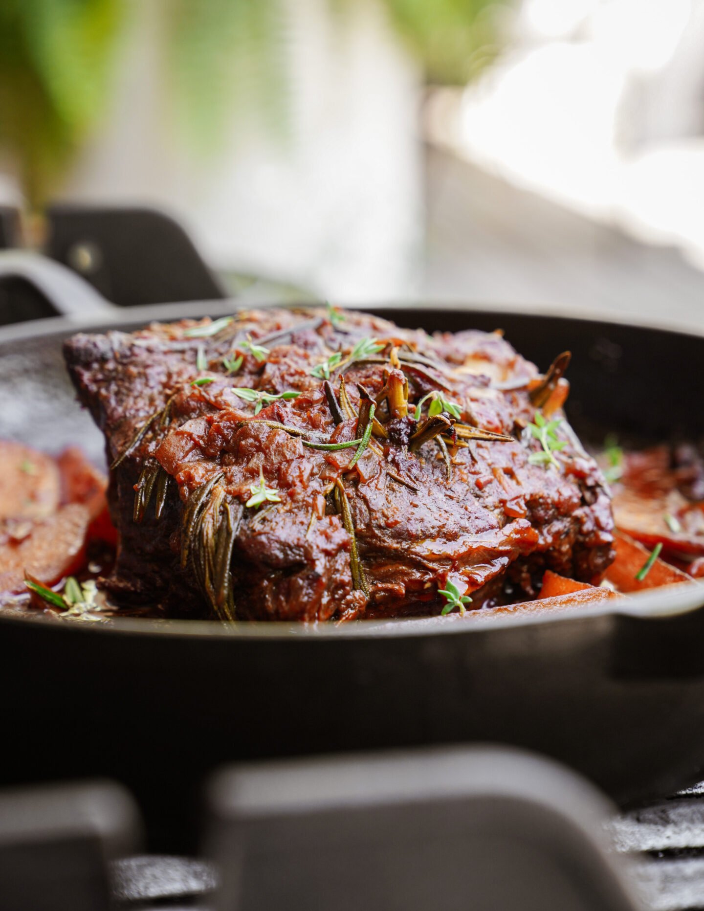 A close-up of a braised beef roast topped with fresh herbs and surrounded by vegetables in a black cast iron skillet.