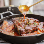 A close-up of a cooked pot roast in a black cast iron pan with carrots and herbs, as a hand pours sauce over the meat using a ladle.