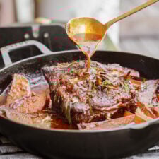 A close-up of a cooked pot roast in a black cast iron pan with carrots and herbs, as a hand pours sauce over the meat using a ladle.