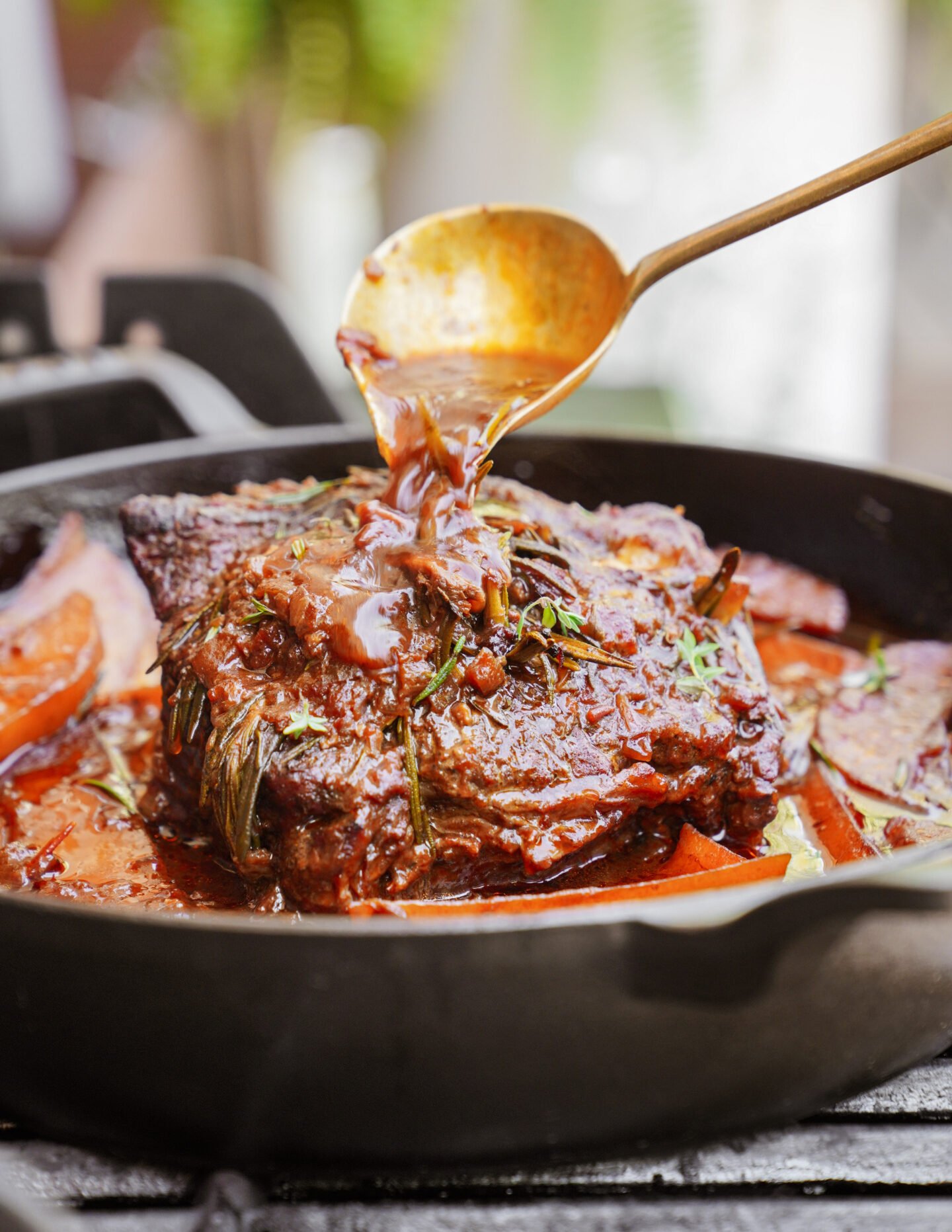 A close-up of a braised beef roast in a cast iron skillet, with a spoon pouring rich brown sauce over the meat. Herbs and sliced vegetables are visible around the roast.