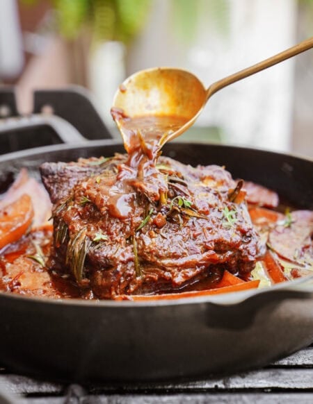 A close-up of a braised beef roast in a cast iron skillet, with a spoon pouring rich brown sauce over the meat. Herbs and sliced vegetables are visible around the roast.
