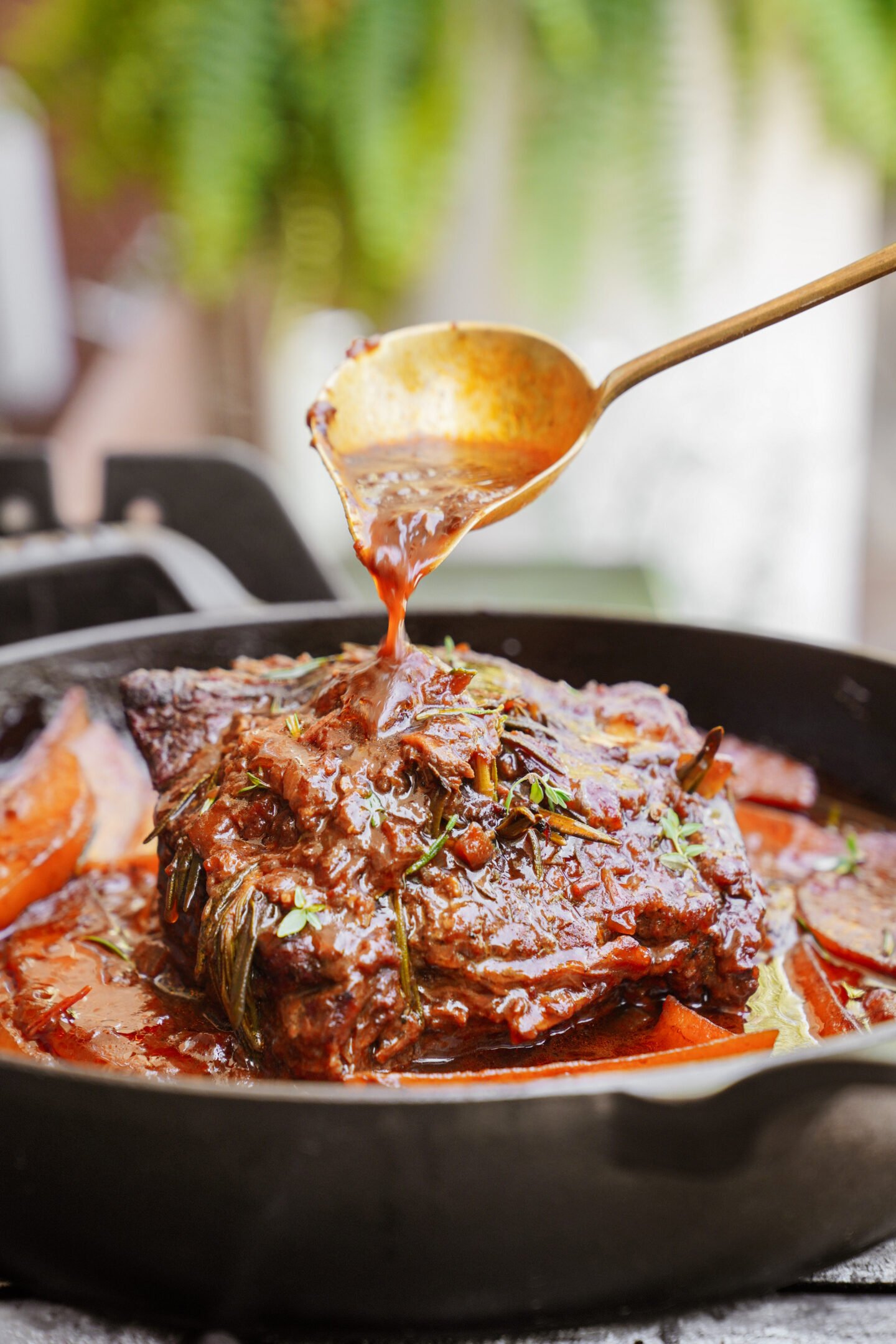 A close-up of a braised beef roast in a cast iron skillet, topped with vegetables and herbs, while a spoon pours savory sauce over the meat.
