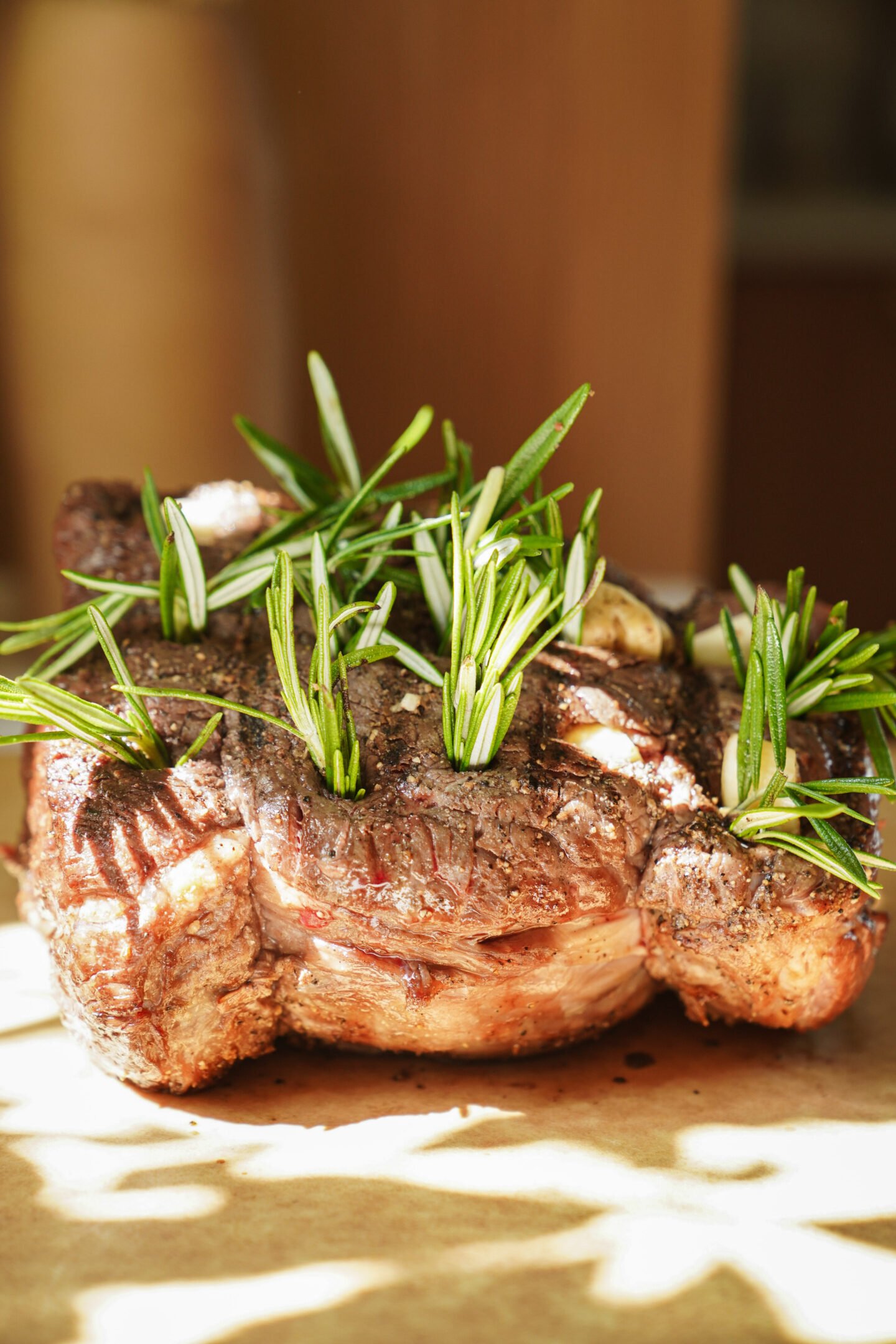 A cooked beef roast seasoned with black pepper and topped with sprigs of fresh rosemary sits on a sheet of parchment paper in warm sunlight.