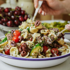 A close-up of a pasta salad with cherry tomatoes, zucchini, olives, and crumbled cheese in a white bowl, with a hand serving some onto a plate. A bowl of cherries is visible in the background.