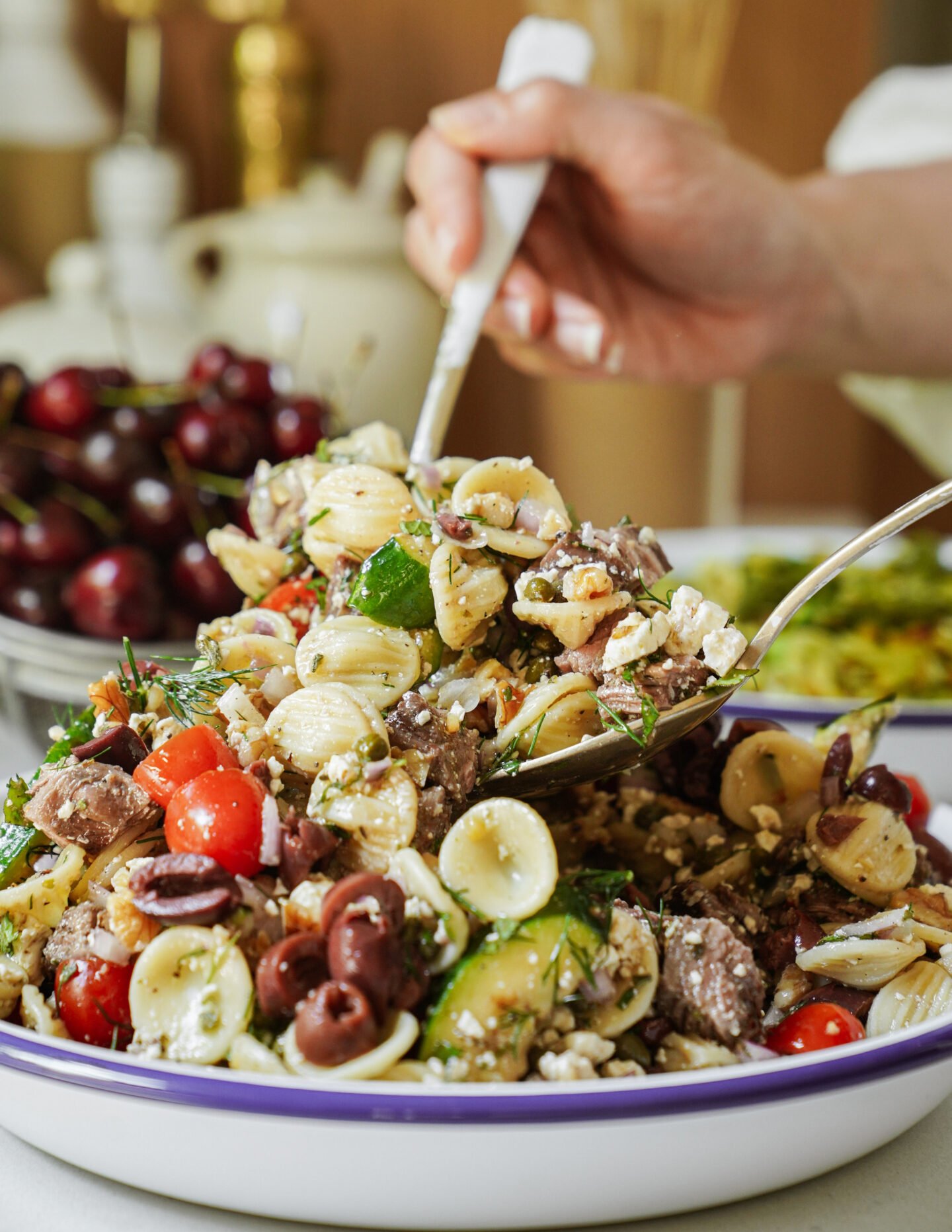 A close-up of a bowl of pasta salad with cherry tomatoes, olives, cucumbers, feta cheese, and herbs. A hand is serving the salad with a fork and spoon. Cherries and another dish are visible in the background.
