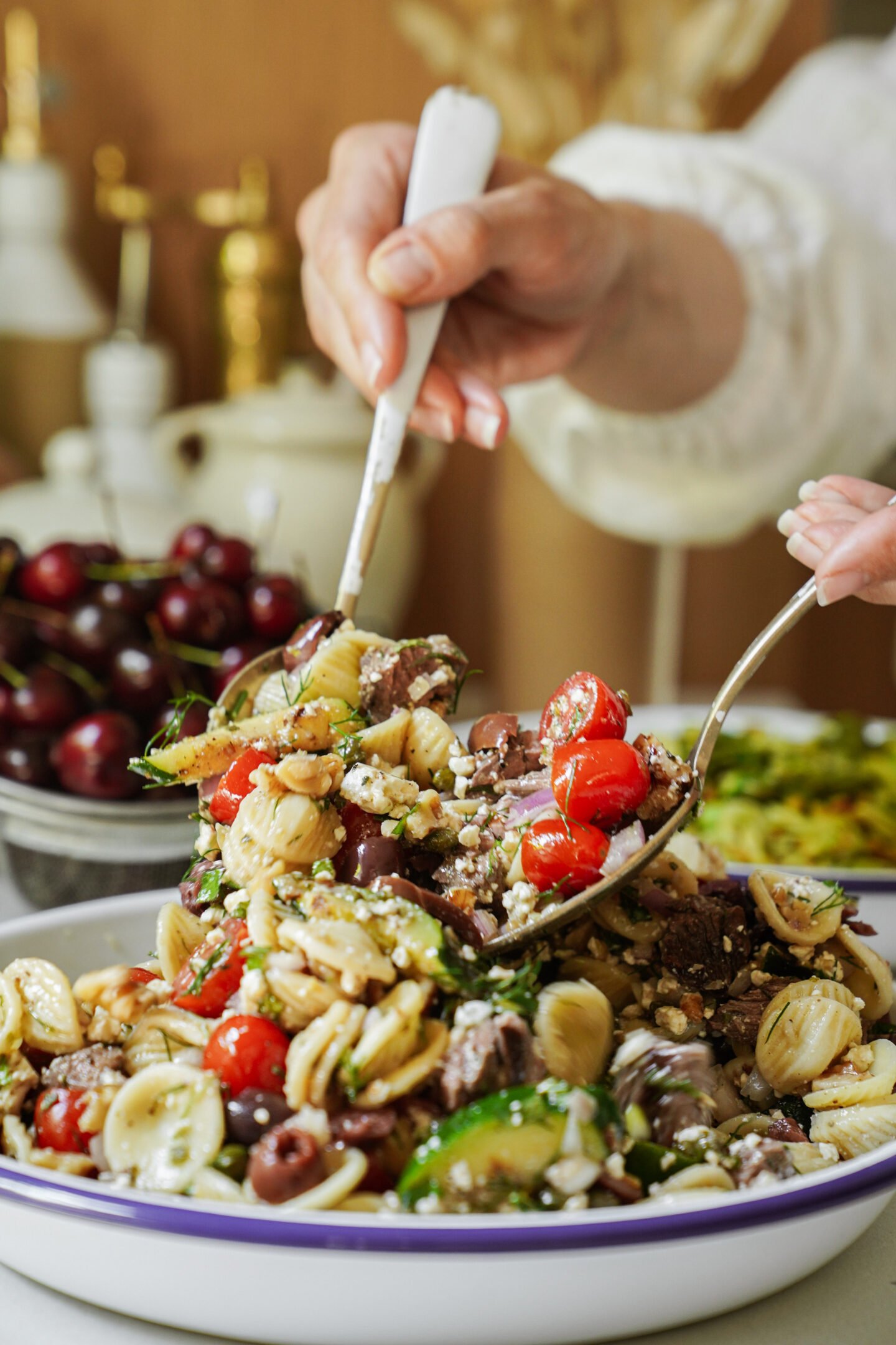 A hand serves a colorful pasta salad with cherry tomatoes, olives, cucumbers, and feta cheese from a large white platter using a spoon and fork. A bowl of cherries and kitchen decor are visible in the background.
