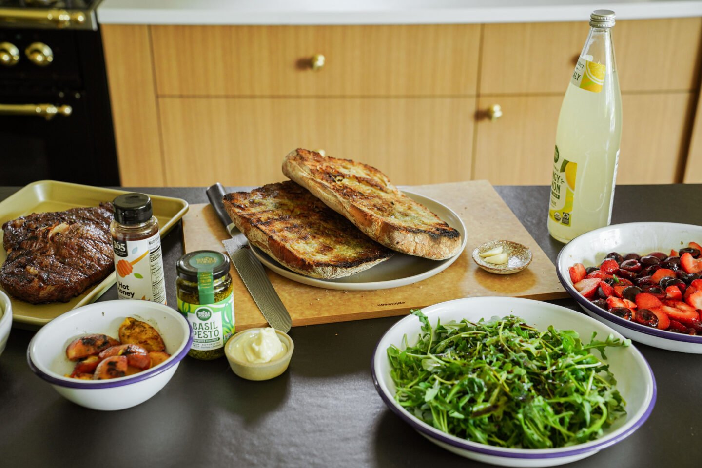 A kitchen counter displays grilled bread, arugula salad, a steak, condiment jars, a bowl of sliced stone fruit, a berry salad, and a bottle of sparkling lemonade, ready for a meal.