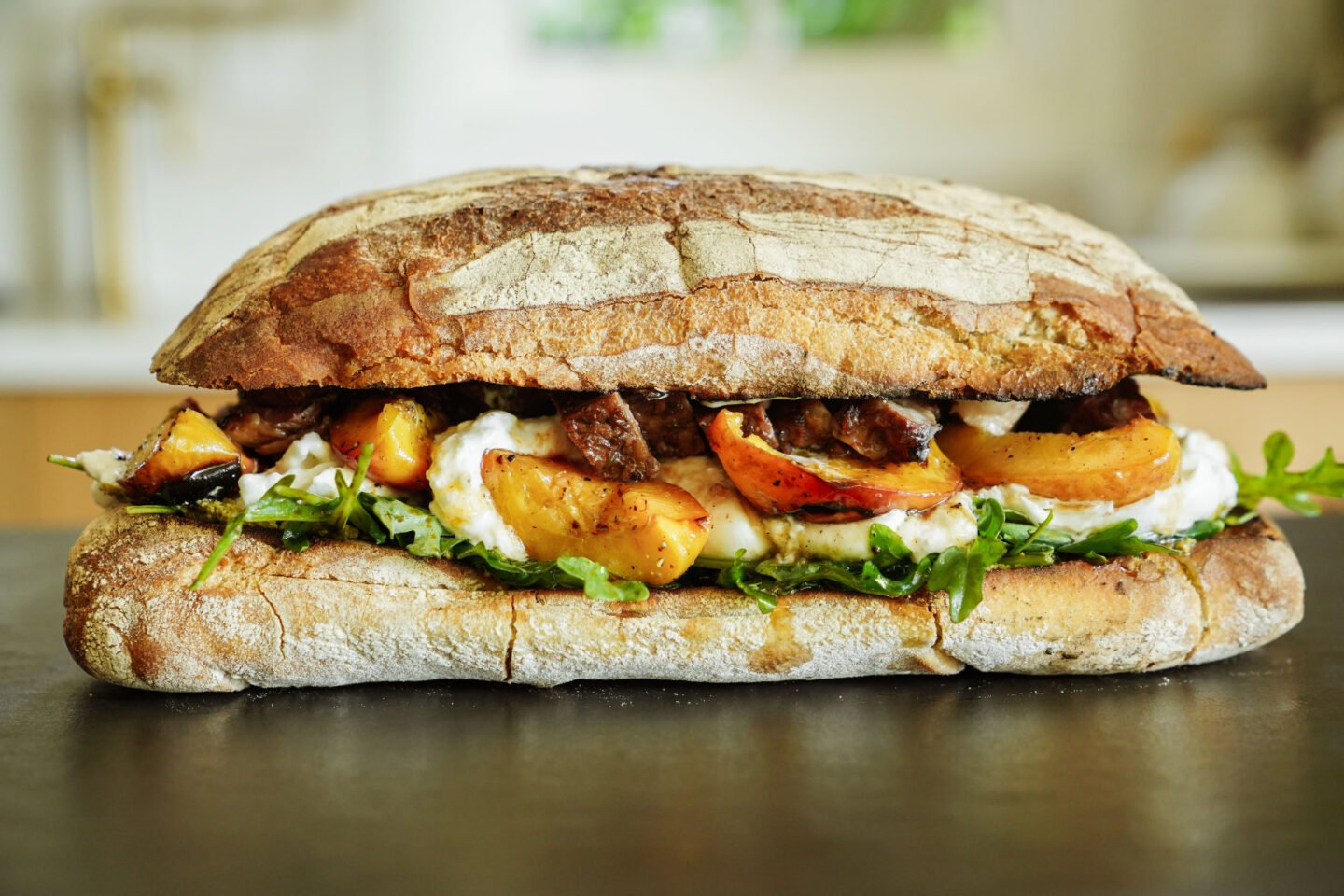 A rustic ciabatta sandwich filled with leafy greens, grilled peaches, creamy cheese, and chunks of roasted meat, set on a dark surface with a blurred kitchen background.
