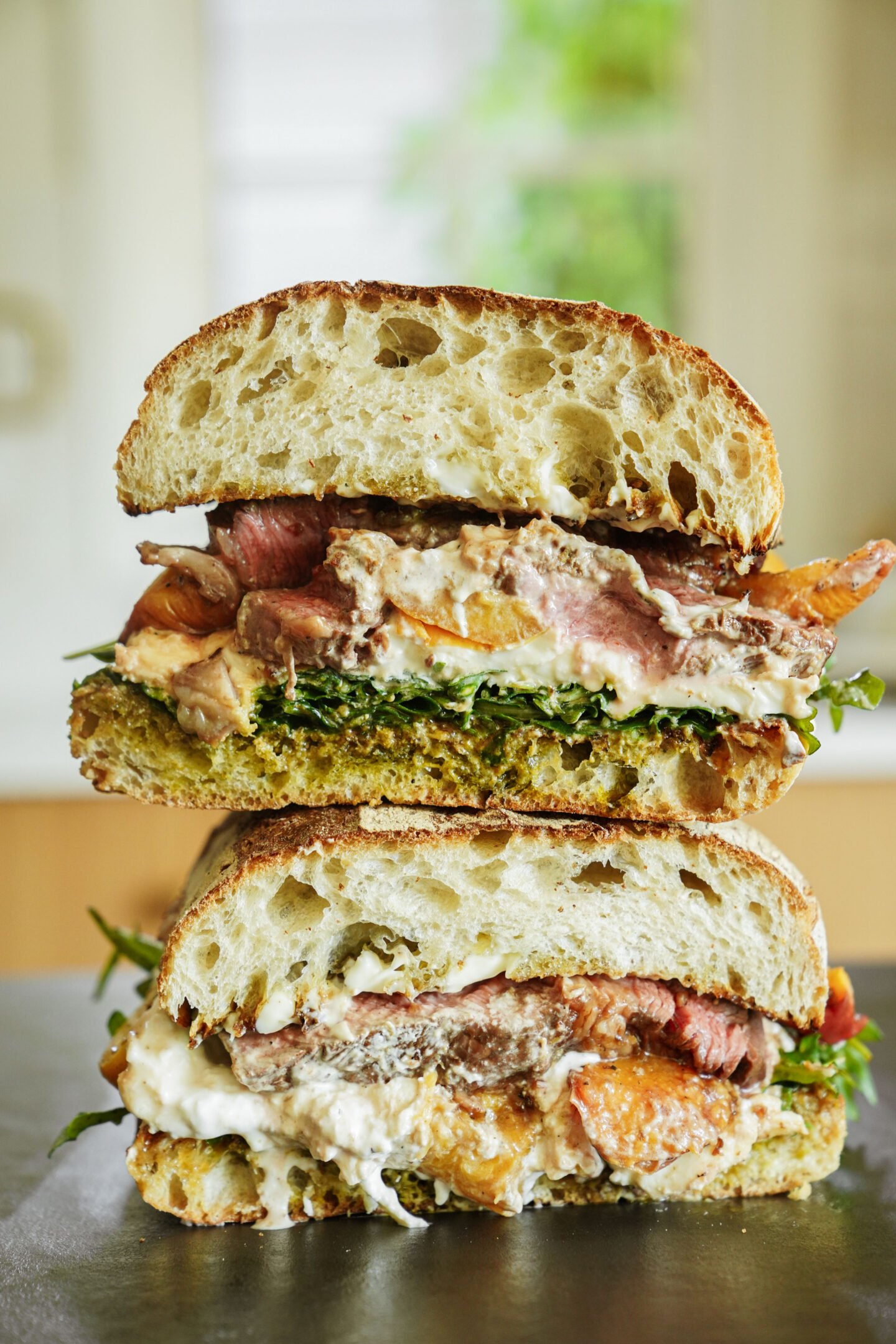 Two stacked halves of a rustic sandwich on ciabatta bread, filled with sliced steak, arugula, pesto, and creamy cheese, set against a softly blurred kitchen background.